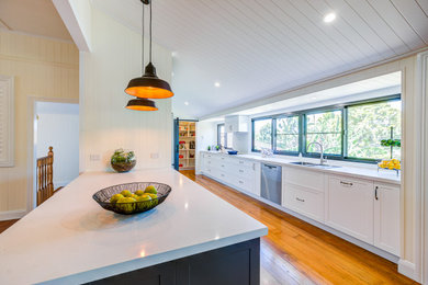 Photo of an expansive contemporary galley eat-in kitchen in Brisbane with a double-bowl sink, shaker cabinets, white cabinets, quartz benchtops, white splashback, engineered quartz splashback, stainless steel appliances, light hardwood floors, with island, brown floor, white benchtop and wood.