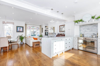 This is an example of a medium sized modern u-shaped open plan kitchen in London with a built-in sink, shaker cabinets, white cabinets, marble worktops, white splashback, marble splashback, integrated appliances, light hardwood flooring, an island, brown floors and white worktops.