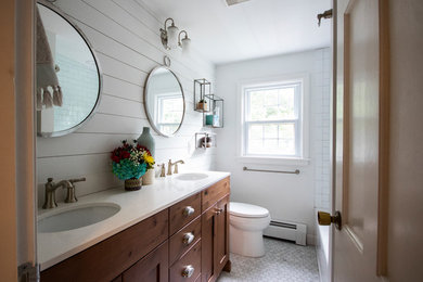 Photo of a mid-sized country master bathroom in Providence with recessed-panel cabinets, medium wood cabinets, an alcove tub, a shower/bathtub combo, a two-piece toilet, white tile, porcelain tile, white walls, mosaic tile floors, an undermount sink, engineered quartz benchtops, white floor, a hinged shower door and white benchtops.