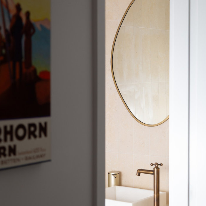 Elegant powder room design featuring warm limestone-look wall tile, a sculptural asymmetrical mirror, and un-lacquered brass fixtures. The minimalist custom vanity and soft neutral palette create a se