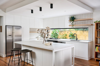 Large contemporary l-shaped open plan kitchen in Perth with an undermount sink, flat-panel cabinets, white cabinets, quartz benchtops, beige splashback, porcelain splashback, black appliances, medium hardwood floors, with island, brown floor and white benchtop.
