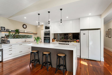 Photo of a contemporary galley open plan kitchen in Melbourne with a double-bowl sink, white cabinets, quartz benchtops, metallic splashback, glass sheet splashback, black appliances, medium hardwood floors, with island, brown floor and white benchtop.