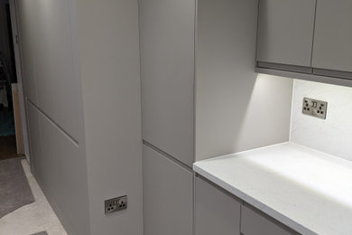 Photo of a medium sized modern grey and white single-wall open plan kitchen in Other with a belfast sink, flat-panel cabinets, grey cabinets, laminate countertops, white splashback, glass sheet splashback, white appliances, carpet, no island, grey floors, white worktops and feature lighting.