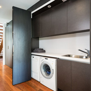 Modelo de armario lavadero lineal, minimalista, de tamaño medio, con fregadero encastrado, encimera de cuarzo compacto, paredes blancas, suelo de madera en tonos medios, lavadora y secadora juntas, armarios con paneles lisos, encimeras blancas y puertas de armario marrones