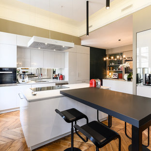 Cette image montre une cuisine design avec un évier encastré, un placard à porte plane, des portes de placard blanches, un électroménager en acier inoxydable, un sol en bois brun, un îlot central, un sol marron et un plan de travail blanc.