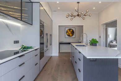 Photo of a mid-sized industrial galley kitchen in Auckland with a double-bowl sink, flat-panel cabinets, grey cabinets, white splashback, coloured appliances, light hardwood floors, with island and white benchtop.