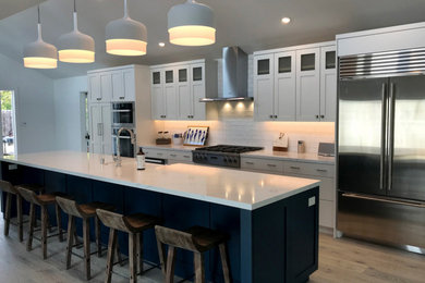 Large trendy single-wall brown floor eat-in kitchen photo in San Francisco with recessed-panel cabinets, white cabinets, white backsplash, stainless steel appliances, an island, white countertops and a drop-in sink