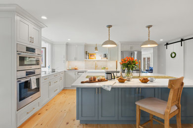 Example of a large country l-shaped light wood floor and beige floor kitchen design in Boston with a farmhouse sink, recessed-panel cabinets, marble countertops, marble backsplash, stainless steel appliances, an island, white countertops, white cabinets and white backsplash