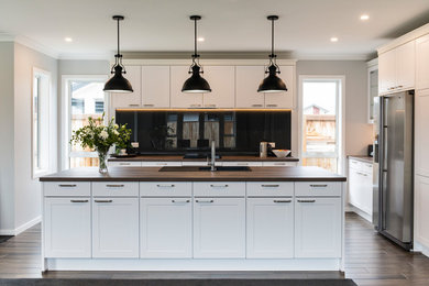Photo of a large transitional l-shaped kitchen pantry in Christchurch with a farmhouse sink, shaker cabinets, white cabinets, laminate benchtops, black splashback, glass sheet splashback, stainless steel appliances, vinyl floors, with island and brown floor.