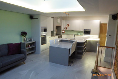 Large contemporary l-shaped open plan kitchen in Kent with a submerged sink, flat-panel cabinets, grey cabinets, quartz worktops, green splashback, glass sheet splashback, stainless steel appliances, porcelain flooring, an island and grey floors.