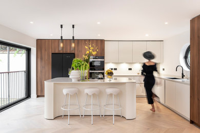 Design ideas for a large contemporary cream and black l-shaped kitchen/diner in London with a built-in sink, flat-panel cabinets, white cabinets, marble worktops, white splashback, marble splashback, black appliances, light hardwood flooring, a breakfast bar and white worktops.