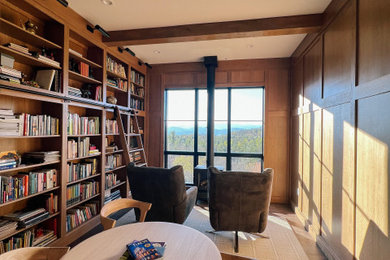 Foto de sala de estar con biblioteca abierta vintage de tamaño medio con paredes marrones, suelo de madera clara, estufa de leña, marco de chimenea de metal, vigas vistas y madera