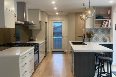 Photo of a mid-sized transitional galley eat-in kitchen in Melbourne with a double-bowl sink, flat-panel cabinets, green cabinets, quartz benchtops, grey splashback, mirror splashback, black appliances, laminate floors, a peninsula, brown floor and white benchtop.