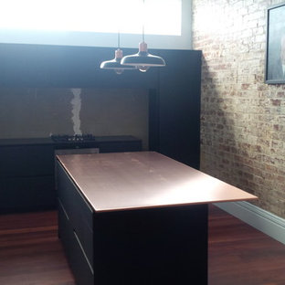 Exemple d'une arrière-cuisine linéaire industrielle de taille moyenne avec un évier 1 bac, des portes de placard noires, un plan de travail en cuivre, une crédence métallisée, une crédence en feuille de verre, un électroménager noir, un sol en bois brun et un îlot central.