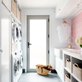 Photo of a medium sized coastal galley separated utility room in Brisbane with a belfast sink, open cabinets, white cabinets, a stacked washer and dryer, grey floors, white walls and white worktops.