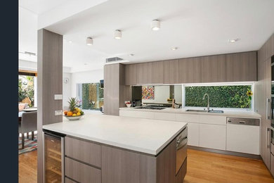 Large galley eat-in kitchen in Sydney with an undermount sink, flat-panel cabinets, light wood cabinets, quartz benchtops, multi-coloured splashback, mirror splashback, stainless steel appliances, light hardwood floors, with island, beige floor and white benchtop.