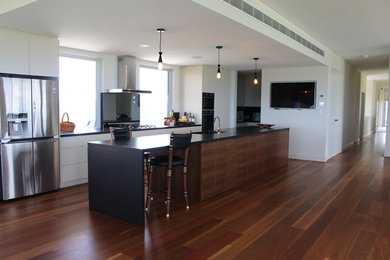 This is an example of an expansive contemporary galley kitchen in Newcastle - Maitland with an undermount sink, flat-panel cabinets, beige cabinets, solid surface benchtops, grey splashback, mirror splashback, stainless steel appliances, medium hardwood floors, with island, multi-coloured floor and black benchtop.