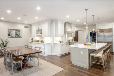 Photo of a large transitional u-shaped eat-in kitchen in Cincinnati with a farmhouse sink, beaded inset cabinets, white cabinets, quartz benchtops, white splashback, ceramic splashback, stainless steel appliances, medium hardwood floors, with island, brown floor and white benchtop.