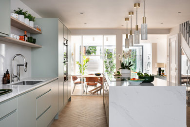 Large contemporary galley open plan kitchen in London with an integrated sink, flat-panel cabinets, green cabinets, quartz worktops, white splashback, engineered quartz splashback, black appliances, ceramic flooring, an island, beige floors, white worktops, all types of ceiling and feature lighting.