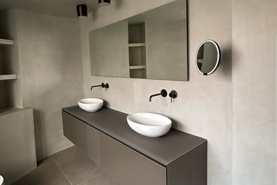Medium sized modern grey and brown ensuite bathroom in London with flat-panel cabinets, grey cabinets, a freestanding bath, a walk-in shower, a wall mounted toilet, ceramic flooring, a console sink, glass worktops, brown floors, an open shower, grey worktops, a feature wall, double sinks and a floating vanity unit.