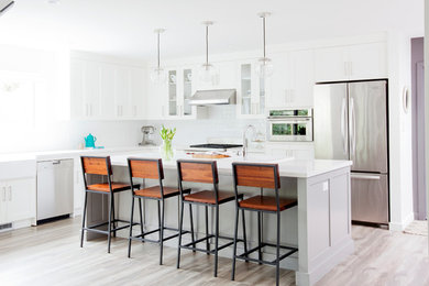 Large transitional l-shaped kitchen in Vancouver with shaker cabinets, white cabinets, solid surface benchtops, white splashback, subway tile splashback, with island, stainless steel appliances and light hardwood floors.