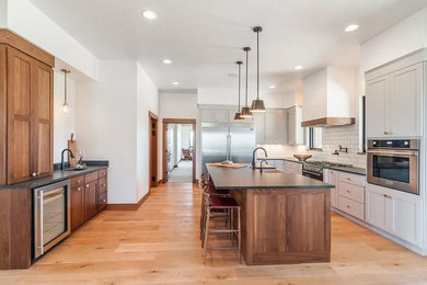 Large trendy l-shaped light wood floor kitchen photo in Other with a farmhouse sink, shaker cabinets, medium tone wood cabinets, granite countertops, white backsplash, subway tile backsplash, stainless steel appliances, an island and black countertops