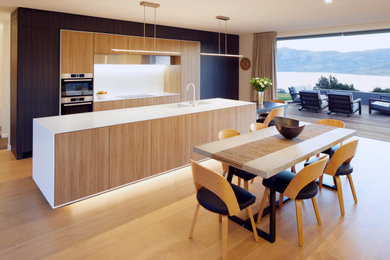 Inspiration for a mid-sized contemporary single-wall eat-in kitchen in Christchurch with an integrated sink, flat-panel cabinets, light wood cabinets, white splashback, light hardwood floors, with island, beige floor, white benchtop and stainless steel appliances.