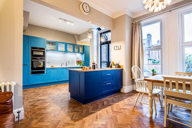 Inspiration for a large contemporary galley eat-in kitchen in Manchester with a farmhouse sink, shaker cabinets, blue cabinets, glass benchtops, white splashback, ceramic splashback, panelled appliances, vinyl floors, with island, brown floor and white benchtop.