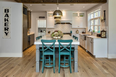 Inspiration for a large arts and crafts u-shaped open plan kitchen in Portland Maine with recessed-panel cabinets, white cabinets, quartz benchtops, white splashback, subway tile splashback, with island, a farmhouse sink, stainless steel appliances and light hardwood floors.