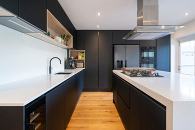 This is an example of a small contemporary l-shaped enclosed kitchen in Oxfordshire with a submerged sink, flat-panel cabinets, black cabinets, quartz worktops, white splashback, engineered quartz splashback, black appliances, light hardwood flooring, an island, brown floors, white worktops and a chimney breast.