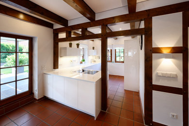 This is an example of a mid-sized country l-shaped open plan kitchen in Essen with an integrated sink, flat-panel cabinets, white cabinets, terra-cotta floors, a peninsula, red floor and white benchtop.