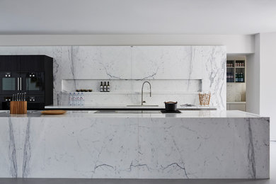 Photo of a mid-sized contemporary galley kitchen in London with an undermount sink, flat-panel cabinets, white cabinets, marble benchtops, with island, grey floor, grey splashback, marble splashback, black appliances and concrete floors.