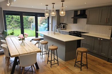Large modern grey and white single-wall kitchen in London with an integrated sink, raised-panel cabinets, grey cabinets, engineered stone countertops, white splashback, ceramic splashback, black appliances, dark hardwood flooring, an island, white worktops, a drop ceiling and feature lighting.