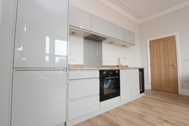 This is an example of a small scandinavian galley open plan kitchen in Sussex with a single-bowl sink, flat-panel cabinets, grey cabinets, wood worktops, grey splashback, glass sheet splashback, black appliances, laminate floors and no island.