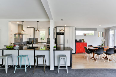 This is an example of a mid-sized modern u-shaped eat-in kitchen in Christchurch with a single-bowl sink, white cabinets, solid surface benchtops, grey splashback, ceramic splashback, stainless steel appliances, medium hardwood floors, multiple islands, beige floor and black benchtop.