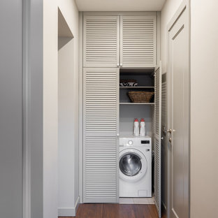 This is an example of a contemporary laundry cupboard in Moscow with louvered cabinets, grey cabinets, dark hardwood flooring, an integrated washer and dryer and brown floors.