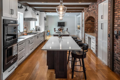 Country galley kitchen/diner in Other with a submerged sink, flat-panel cabinets, medium wood cabinets, engineered stone countertops, white splashback, metro tiled splashback, black appliances, light hardwood flooring, multiple islands, beige floors, white worktops and exposed beams.