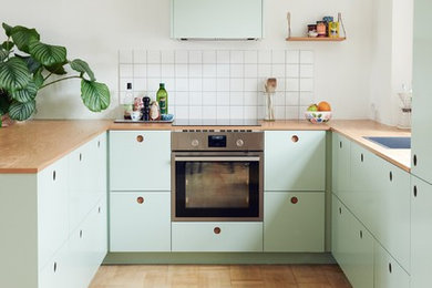 This is an example of a scandinavian kitchen in Copenhagen with a drop-in sink, flat-panel cabinets, green cabinets, wood benchtops, white splashback, stainless steel appliances, medium hardwood floors and a peninsula.