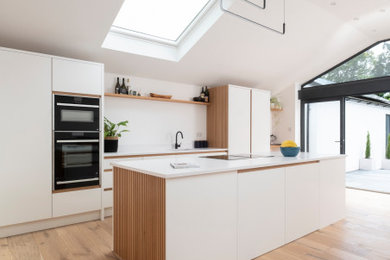 Photo of a medium sized scandi single-wall open plan kitchen in Cardiff with an integrated sink, flat-panel cabinets, white cabinets, quartz worktops, white splashback, stainless steel appliances, light hardwood flooring, an island, white worktops, a vaulted ceiling and feature lighting.