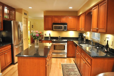 Mid-sized elegant u-shaped light wood floor and beige floor enclosed kitchen photo in Baltimore with a double-bowl sink, raised-panel cabinets, dark wood cabinets, granite countertops, black backsplash, stainless steel appliances and an island