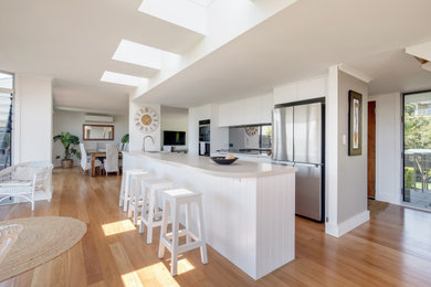 Mid-sized beach style galley open plan kitchen in Sydney with an undermount sink, flat-panel cabinets, white cabinets, quartz benchtops, metallic splashback, mirror splashback, stainless steel appliances, medium hardwood floors, with island and beige benchtop.