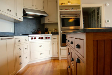 Example of a large classic u-shaped light wood floor and brown floor enclosed kitchen design in Other with white cabinets, an island, a farmhouse sink, shaker cabinets, soapstone countertops, black backsplash, stone slab backsplash, stainless steel appliances and black countertops