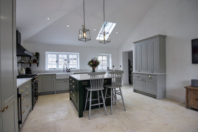 This is an example of a medium sized traditional grey and teal l-shaped kitchen/diner in Buckinghamshire with a belfast sink, shaker cabinets, green cabinets, quartz worktops, metallic splashback, mirror splashback, black appliances, limestone flooring, an island, beige floors, white worktops and a vaulted ceiling.