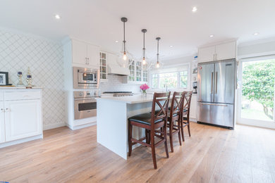 Mid-sized cottage l-shaped light wood floor open concept kitchen photo in Vancouver with white backsplash, stainless steel appliances, an island and white cabinets