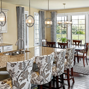 Modelo de cocina comedor de galera, tradicional, de tamaño medio, con fregadero bajoencimera, armarios con paneles con relieve, puertas de armario blancas, salpicadero beige, suelo de madera oscura, una isla y salpicadero de azulejos de piedra