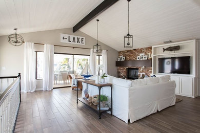 Example of a mid-sized beach style loft-style medium tone wood floor and brown floor living room design in Los Angeles with beige walls, a standard fireplace, a brick fireplace and a wall-mounted tv
