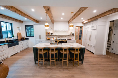 This is an example of a large eclectic l-shaped open plan kitchen in Toronto with a farmhouse sink, quartz benchtops, white splashback, engineered quartz splashback, black appliances, light hardwood floors, with island and white benchtop.