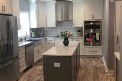Photo of a mid-sized traditional l-shaped separate kitchen in Baltimore with a farmhouse sink, shaker cabinets, white cabinets, white splashback, stainless steel appliances, light hardwood floors, with island, brown floor, solid surface benchtops and subway tile splashback.