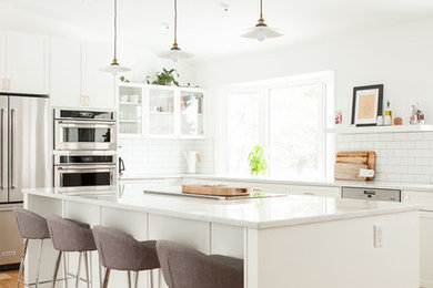 Transitional l-shaped light wood floor kitchen photo in Other with glass-front cabinets, white cabinets, white backsplash, subway tile backsplash, stainless steel appliances and an island
