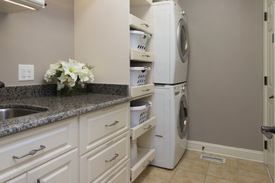 Mid-sized elegant single-wall porcelain tile and beige floor dedicated laundry room photo in Raleigh with raised-panel cabinets, white cabinets, granite countertops, a stacked washer/dryer, an undermount sink and gray walls